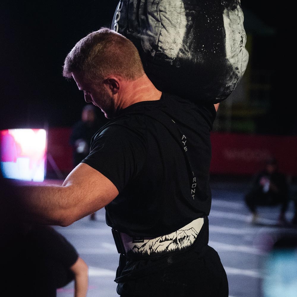 Athlet beim Strongman-Event hebt einen schweren Sandsack über die Schulter und trägt den 2POOD Summit Gewichthebergürtel mit Bergmotiv; dunkle Wettkampfszene mit Spotlights.